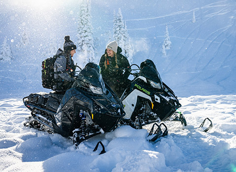 Two people on Polaris snowmobiles, one black and one black and white, are stopped in deep snow, surrounded by snow-covered trees and glistening snow.