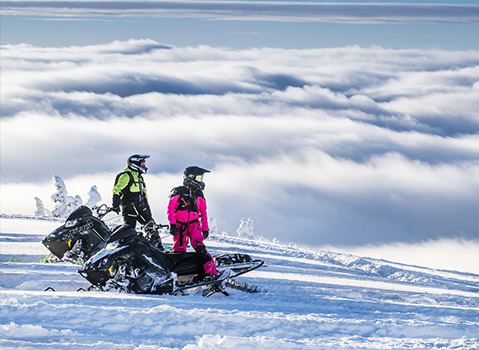 Two snowmobilers in brightly colored gear stand beside their Polaris snowmobiles on a snowy mountain ridge overlooking a vast expanse of clouds.
