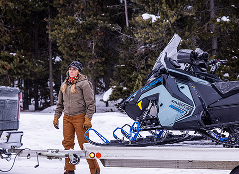 A woman stands next to a Polaris Adventure 550 snowmobile loaded onto a trailer in a snowy forest.