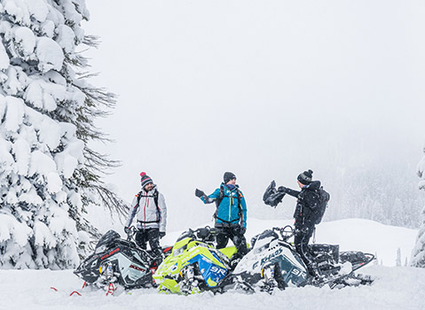 Three people stand amidst several snowmobiles, two of which are overturned, in a heavily snow-laden forest clearing.