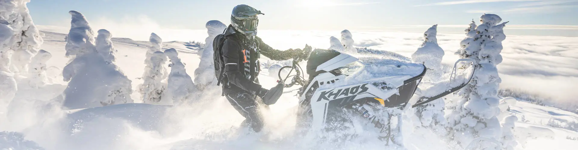 A person in a helmet and blue winter gear rides a white and purple Polaris Snowmobile RMK through deep snow in a forest.
