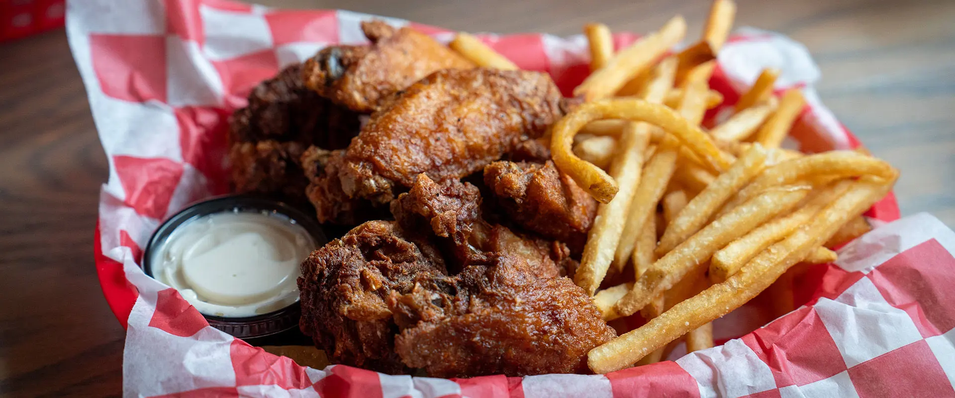 Basket of crispy fried chicken wings, golden French fries, and a side of creamy dipping sauce on a red and white checkered liner.