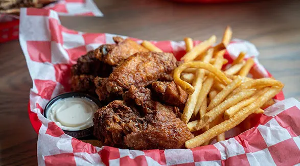 Basket of crispy fried chicken wings, golden French fries, and a side of creamy dipping sauce on a red and white checkered liner.