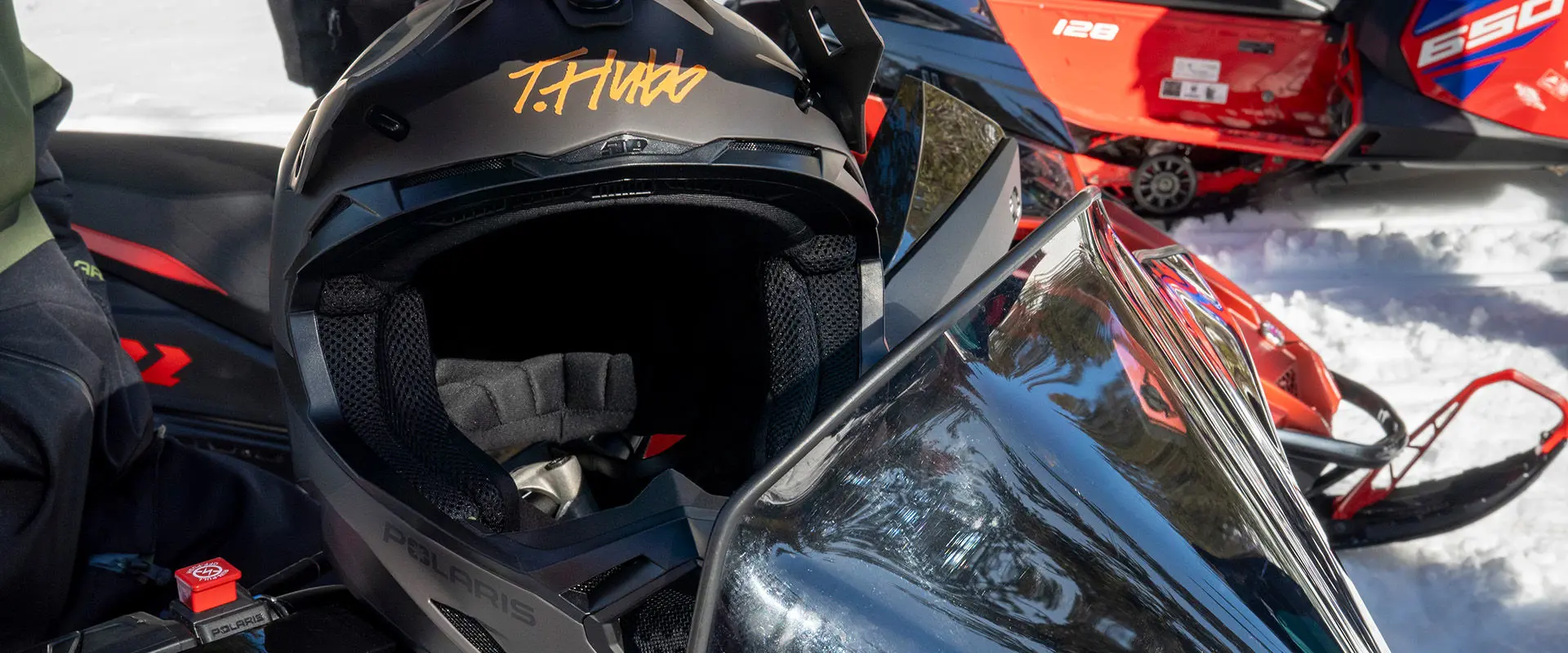 Black Polaris snowmobile helmet featuring a signature graphic "T.Hundo", resting on a Polaris Snowmobile in a snowy environment.