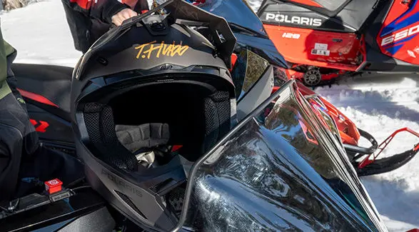 Black Polaris snowmobile helmet featuring a signature graphic "T.Hundo", resting on a Polaris Snowmobile in a snowy environment.