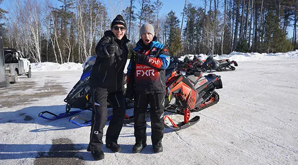 Levi LaVallee, wearing Red Bull and FXR gear, and Luke Gulbranson pose with Polaris snowmobiles on a snowy, tree-lined landscape.