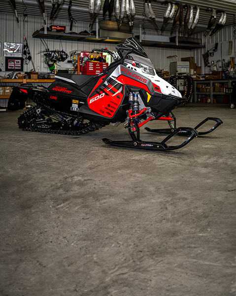 A red, white, and black Polaris 600 INDY Cross Country snowmobile is parked indoors in a well-equipped garage or workshop setting.