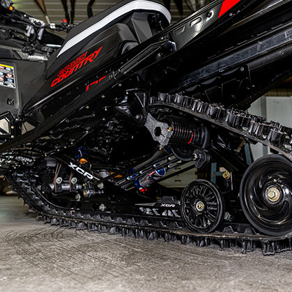 A side view of a red and black snowmobile with the text ‘Cross Country’ visible on its side panel. The snowmobile is parked indoors, with a sign in the background that reads ‘Woody’s 2016 Soo I-500.