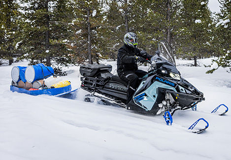 Rider on a Polaris snowmobile pulling a blue utility sled loaded with equipment through a snowy forest.