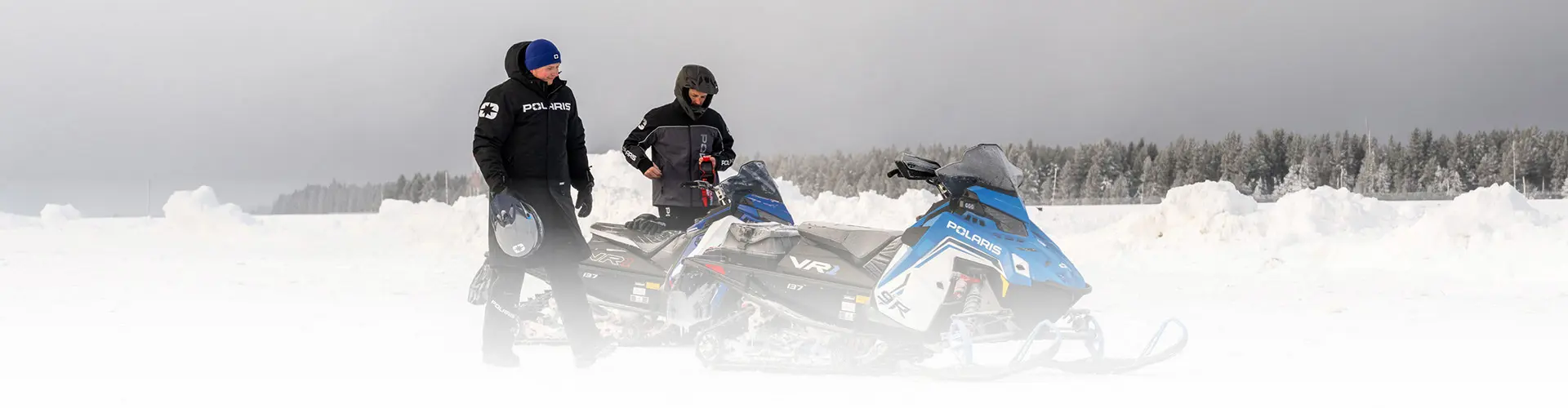 Two riders preparing blue Polaris INDY VR1 snowmobiles on a snow-covered trail near a forest.