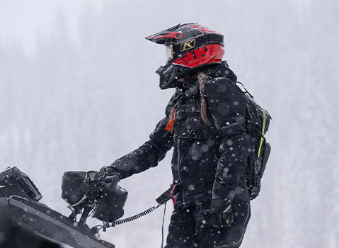Helmeted snowmobile rider wearing Polaris winter gear beside a snowmobile during heavy snowfall.