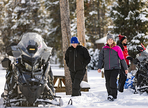 Three riders walking through deep snow beside two Polaris snowmobiles at a wooded winter cabin.