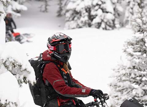 Helmeted rider in red Polaris snow gear riding through deep snow among snow-covered pine trees.