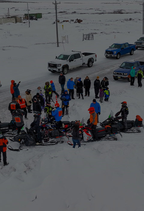 Polaris snowmobile riders and event staff gathered with touring snowmobiles at a snowy outdoor checkpoint.