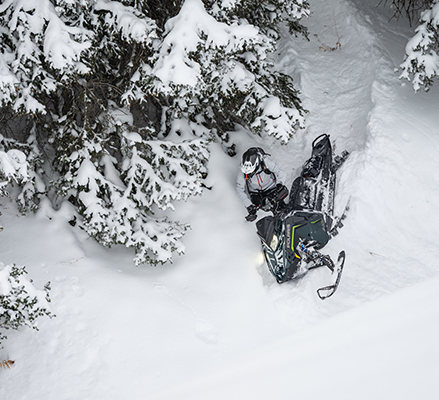 High-angle shot of a rider with a black and fluorescent green details Polaris snowmobile on a steep, snow-covered slope near evergreen trees.