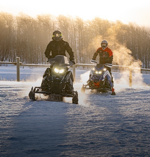 Two Polaris snowmobiles driving on a snowy roadside at sunrise, with riders wearing helmets and winter gear as exhaust vapor rises in the cold air.
