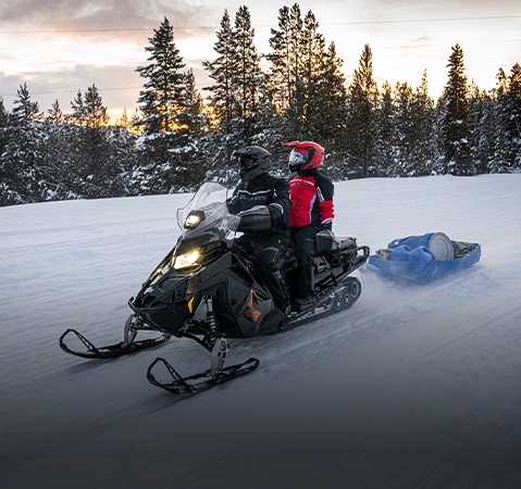 Polaris snowmobile towing a cargo sled across a snowy trail, carrying two riders through a winter landscape lined with snow-covered pine trees.