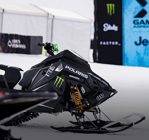 Close-up of a Polaris snowmobile displayed at an indoor event booth, featuring Monster Energy branding, performance suspension components, and sponsor decals.