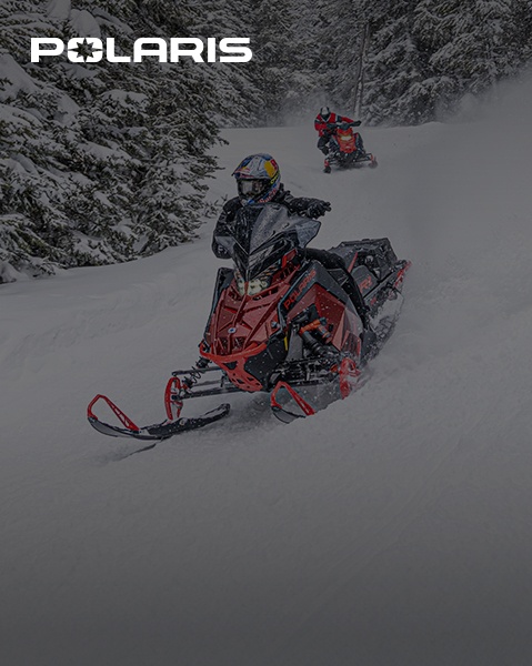 Two people ride Polaris snowmobiles, one red and one grey, through a snow-covered forest, with the Polaris Adventures logo in the upper left corner.