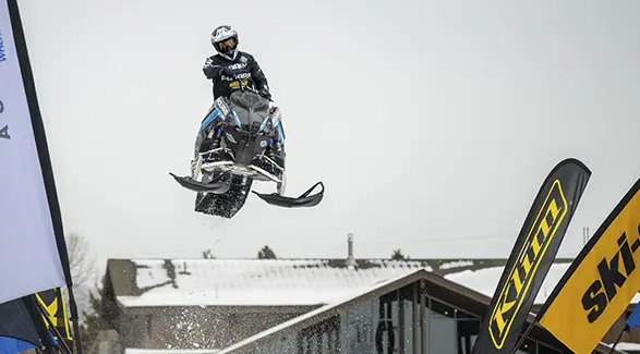 Andy Thomas jumping a black and blue Polaris snowmobile over a snowy event course.