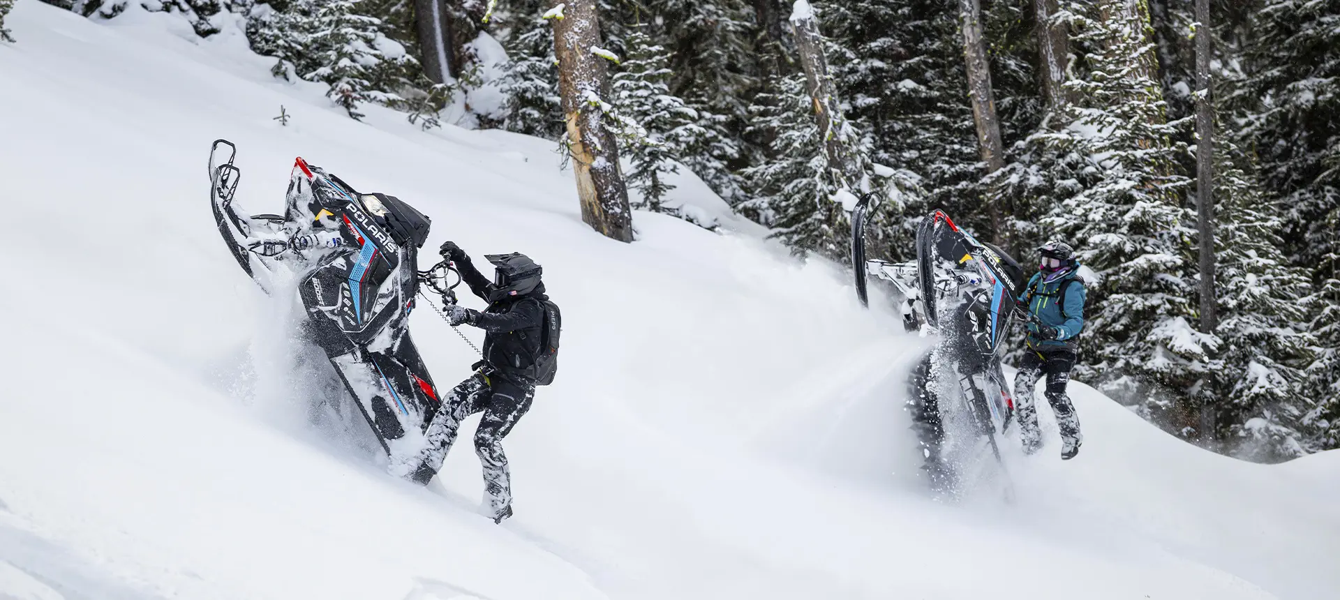 Andy Thomas and another helmeted rider carving Polaris RMK snowmobiles through deep powder in a snow-covered forest.