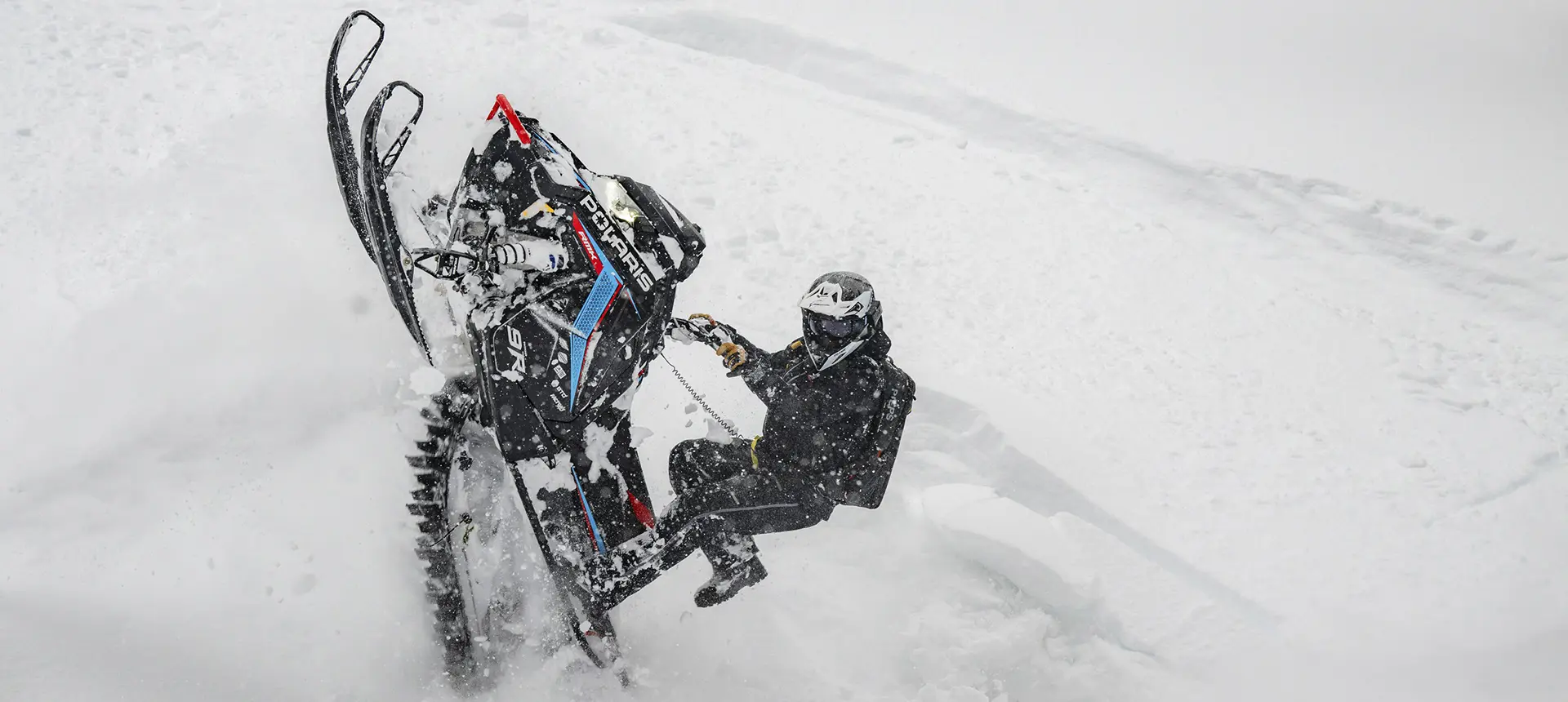 Andy Thomas lifting a Polaris RMK snowmobile through deep powder on a steep snowy slope.