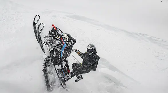 Andy Thomas lifting a Polaris RMK snowmobile through deep powder on a steep snowy slope.