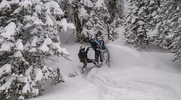 Andy Thomas maneuvering a Polaris RMK snowmobile through deep powder in a snow-covered evergreen forest.