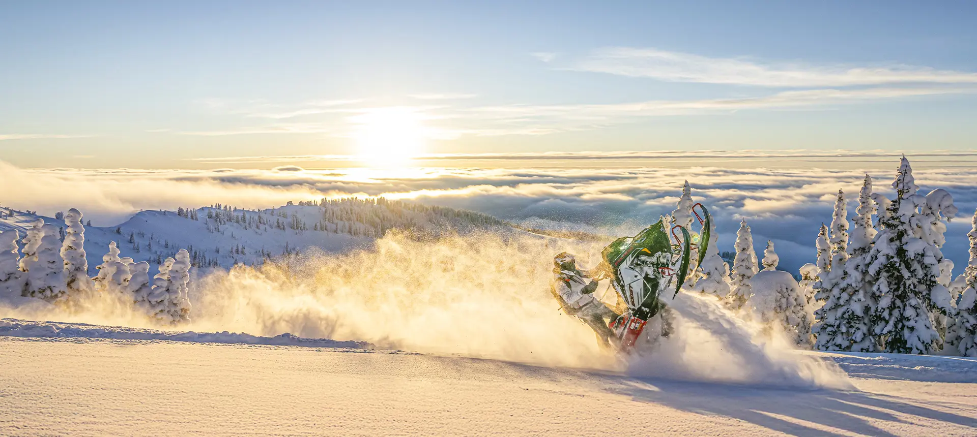 Polaris snowmobile Andy Thomas rider carving through mountain powder at sunrise above snow-covered trees and clouds.