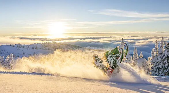 Polaris snowmobile Andy Thomas rider carving through mountain powder at sunrise above snow-covered trees and clouds.