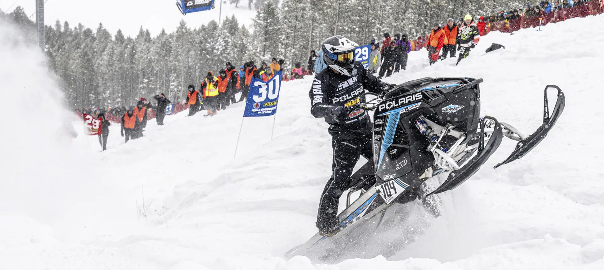 Andy Thomas racing a black Polaris snowmobile uphill as spectators watch from the snowy course.