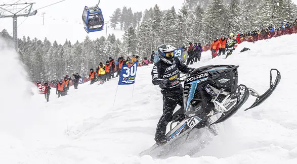 Andy Thomas racing a black Polaris snowmobile uphill as spectators watch from the snowy course.