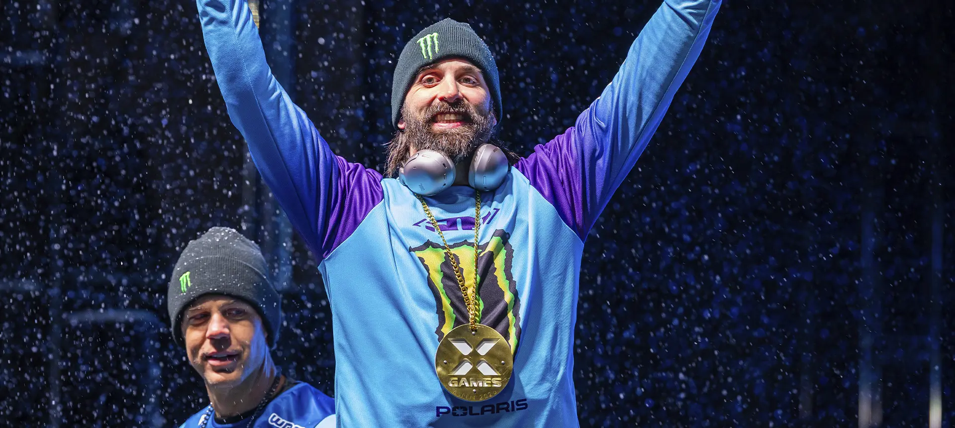 Brett Turcotte raising both arms while wearing a gold X Games medal during snowfall.