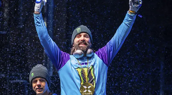 Brett Turcotte raising both arms while wearing a gold X Games medal during snowfall.