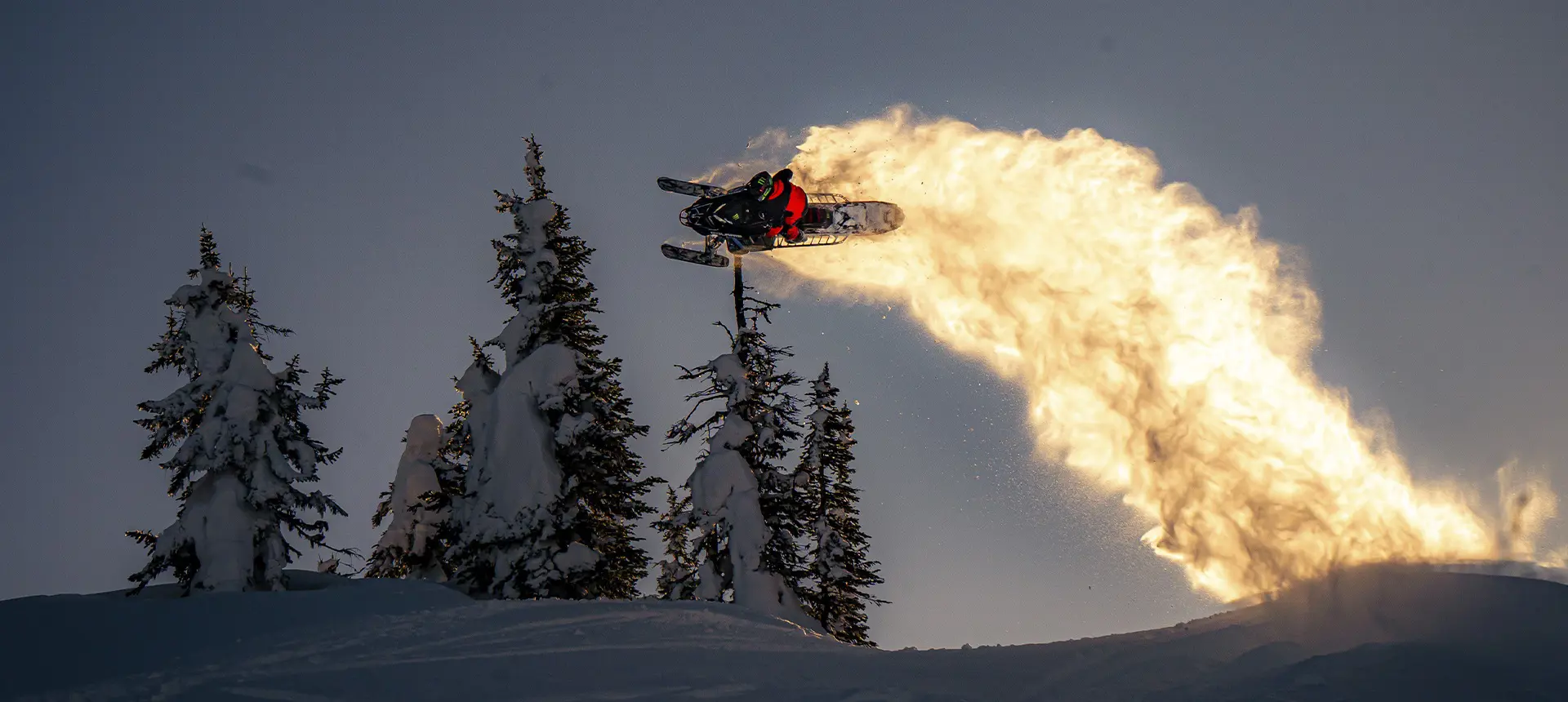 Brett Turcotte launching a Polaris snowmobile above snow-covered trees with a golden trail of powder.