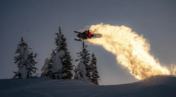 Brett Turcotte launching a Polaris snowmobile above snow-covered trees with a golden trail of powder.