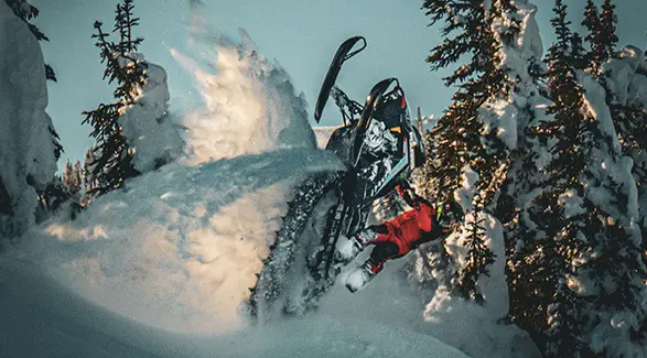 Brett Turcotte carving a black Polaris snowmobile through deep powder in a snow-covered forest.