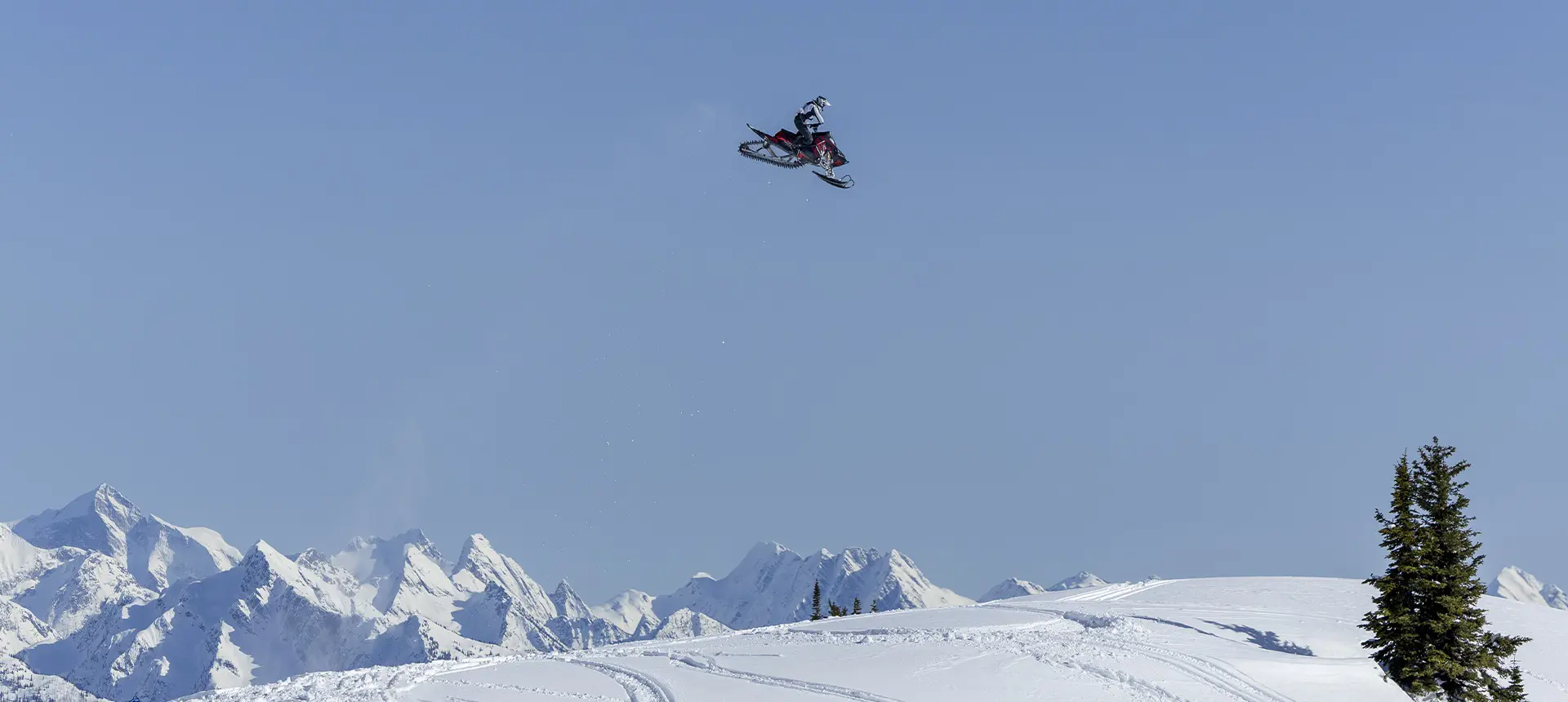 Caleb Kesterke launching a Polaris snowmobile high above a snowy ridge with rugged mountain peaks in the distance.