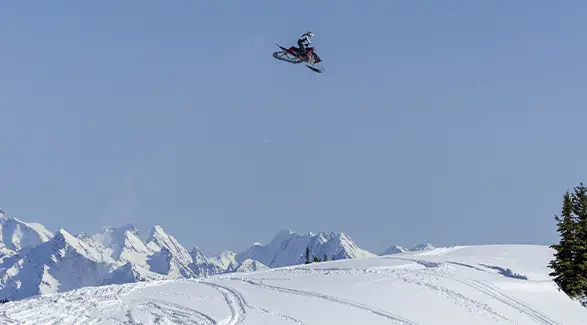 Caleb Kesterke launching a Polaris snowmobile high above a snowy ridge with rugged mountain peaks in the distance.