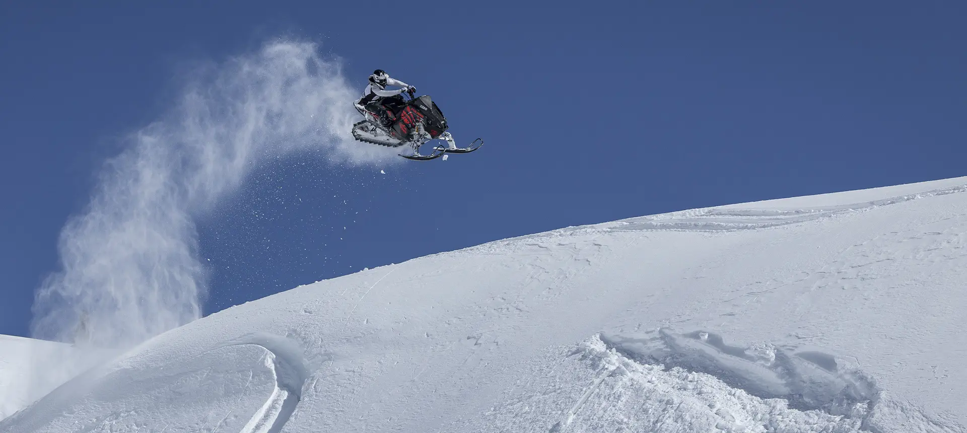 Caleb Kesterke launching a red Polaris RMK snowmobile above a snowy ridge under a clear blue sky.