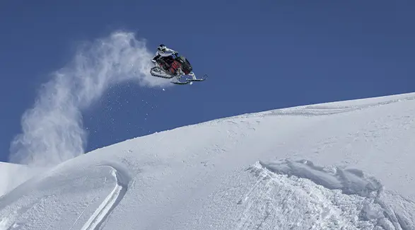Caleb Kesterke launching a red Polaris RMK snowmobile above a snowy ridge under a clear blue sky.