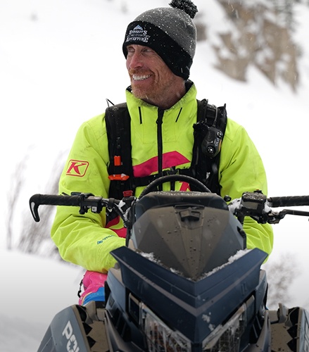 Chris Burandt wearing high-visibility winter gear while seated on a snowmobile in the mountains.