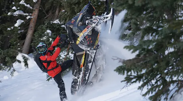 Chris Burandt maneuvering a black Polaris snowmobile through deep powder in a snow-covered evergreen forest.