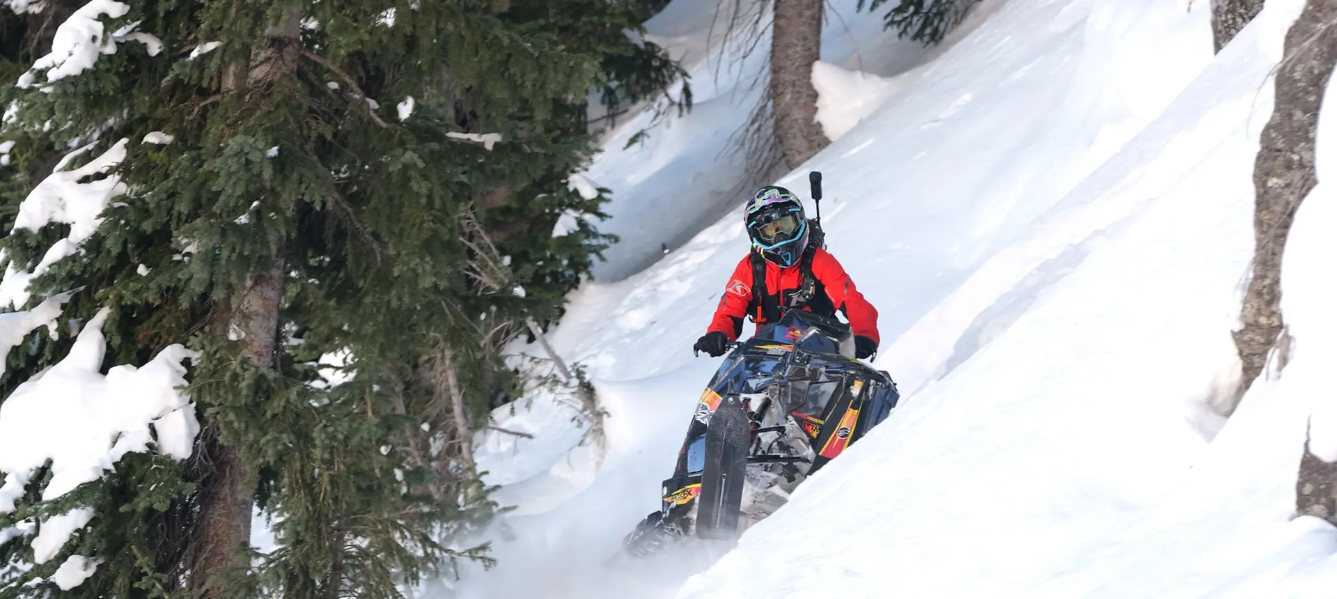 Chris Burandt guiding a black Polaris snowmobile across a steep, tree-lined slope covered in deep snow.