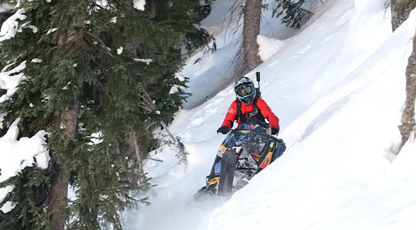 Chris Burandt guiding a black Polaris snowmobile across a steep, tree-lined slope covered in deep snow.