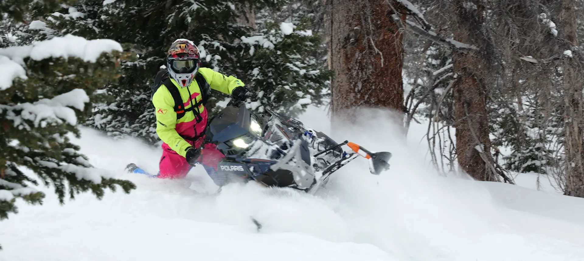 Chris Burandt in high-visibility winter gear carving a Polaris snowmobile through deep powder in a snowy forest.