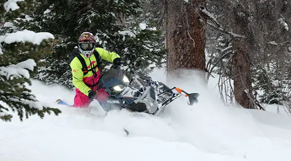 Chris Burandt in high-visibility winter gear carving a Polaris snowmobile through deep powder in a snowy forest.