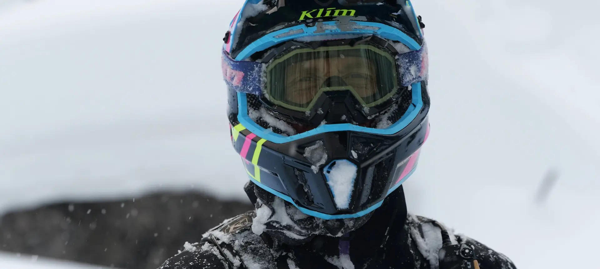 Close-up of a snow-covered snowmobile helmet with colorful blue, pink and neon yellow accents.