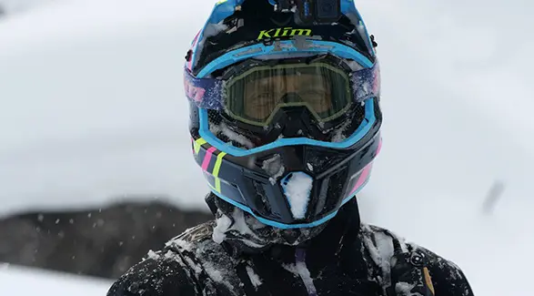 Close-up of a snow-covered snowmobile helmet with colorful blue, pink and neon yellow accents.