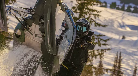 Kyle Pulsifer maneuvering a blue Polaris snowmobile through deep powder on a sunlit mountain slope.